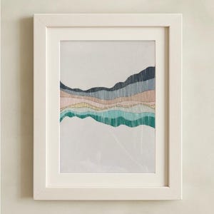 May include: A framed art print featuring an abstract landscape design. The artwork showcases layered horizontal bands in shades of teal, pink, blue, and gold, creating a textured, mountain-like effect. The white frame complements the artwork.