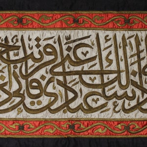 May include: A white banner with gold embroidery of Arabic script, framed by a red and gold border with floral designs. The banner is on a black background.