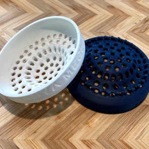 May include: Two 3D-printed objects: a white, oval-shaped bowl and a navy blue, dome-shaped object. Both have a perforated design. The white bowl has the text "YA MEGA" on its side. They sit on a wooden surface with a chevron pattern.