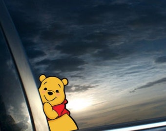 Może przedstawiać: Żółta naklejka z postacią kreskówkową Winnie the Pooh na czarnym oknie samochodu. Naklejka przedstawia Winnie the Pooh zaglądającego zza ramki okna.