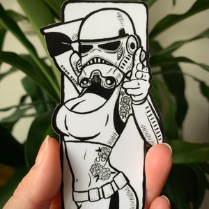 Peut inclure: Autocollant noir et blanc représentant une illustration stylisée d'une Stormtrooper féminine avec des tatouages floraux. Le texte "DADA ZZZ" est présent sur l'autocollant, qui est tenu dans une main.