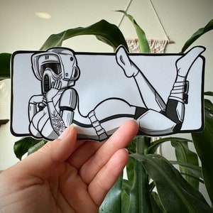 Peut inclure: Une illustration en noir et blanc d'un personnage portant un casque et une combinaison spatiale, allongé sur le dos, les jambes en l'air. Le personnage a un tatouage sur le bras.