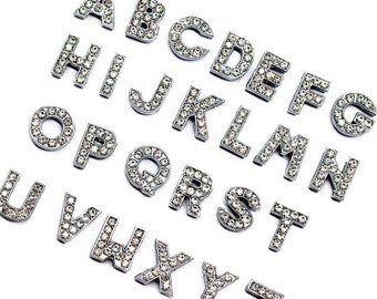 May include: A set of 26 silver-toned alphabet letter charms, each adorned with clear rhinestones. The letters are arranged in alphabetical order.