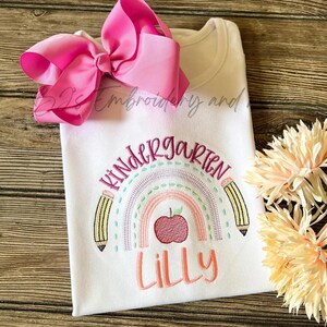 May include: White t-shirt with a rainbow design and the text "Kindergarten" above a red apple and the name "Lilly" below. The shirt is decorated with a pink bow.
