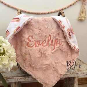 May include: A soft, pink baby blanket with the name "Evelyn" embroidered in a matching color. The blanket has a plush, textured surface and a white border with a rainbow design. The blanket is folded over a basket.