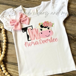 May include: White short-sleeved shirt with pink and black gingham bow, embroidered with a pink cow with flowers, the number "TWO" in pink and black gingham, and the name "Nora Everlee".