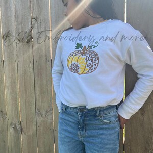 May include: A white long-sleeve shirt featuring an embroidered pumpkin design with yellow sunflower petals and a brown leopard print pattern. The shirt is paired with blue jeans. The text "BJS Embroidery and more" is visible.