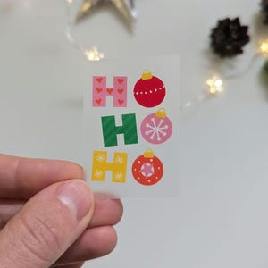 Könnte beinhalten: Ein kleiner Aufkleber mit den Worten "HO HO" in bunten Buchstaben und Weihnachtsschmuck-Designs. Die Buchstaben sind rosa mit Herzen, grün und gelb mit Sternen. Die Ornamente sind rot, rosa und orange.