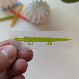 Könnte beinhalten: Ein transparenter Aufkleber mit einer grünen Alligator-Illustration. Der Alligator hat ein weißes Auge und ein kleines Lächeln.