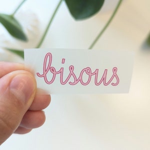 Könnte beinhalten: Eine weiße Karte mit dem Wort "bisous" in rosa Kursivschrift.