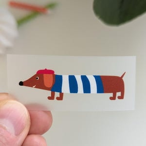 Könnte beinhalten: Rechteckiger Aufkleber mit einem Cartoon-Dackel, der eine rote Baskenmütze und ein blau-weiß gestreiftes Oberteil trägt. Der Hund ist braun mit schwarzer Nase und Augen. Der Aufkleber wird von einer Hand gehalten.