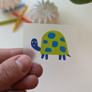 Könnte beinhalten: Ein kleiner, rechteckiger Aufkleber mit einer Cartoon-Schildkröte. Die Schildkröte hat einen blauen Kopf und Gliedmaßen, einen grünen Panzer mit blauen Punkten und ein lächelndes Gesicht. Der Aufkleber wird in einer Hand gehalten.