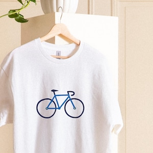 Könnte beinhalten: Weißes Sweatshirt mit blauem Fahrradmotiv. Das Fahrrad-Design hat einen minimalistischen Stil mit blauem Rahmen, Rädern und Lenker. Das Sweatshirt hängt an einem Holzbügel.