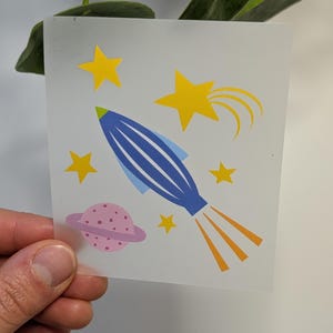 Könnte beinhalten: Eine blaue Rakete mit gelben Flammen und einem rosafarbenen Planeten mit einem Ring. Es gibt fünf gelbe Sterne, von denen einer einen gelben Schweif hat.