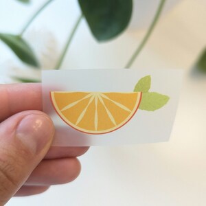 Könnte beinhalten: Ein transparenter Aufkleber mit einer gelben Orangenscheibe und einem grünen Blatt auf weißem Hintergrund.