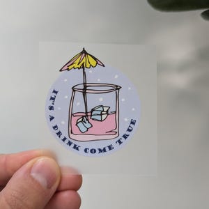 Könnte beinhalten: Ein klarer Aufkleber mit einer Cocktail-Illustration. Das Getränk befindet sich in einem Glas mit Eiswürfeln und einem gelb-rosa Schirm. Der Text "It's a drink come true" ist am Rand des Aufklebers aufgedruckt.