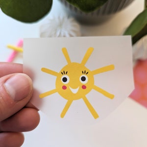 Könnte beinhalten: Eine gelbe Sonne mit einem lächelnden Gesicht und schwarzen Augen. Die Sonne hat rote Wangen und befindet sich auf einem weißen Hintergrund.