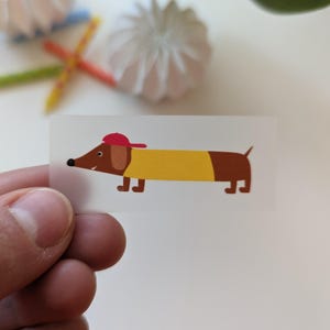 Könnte beinhalten: Ein Aufkleber mit einem Cartoon-Dackel, der ein gelbes Hemd und eine rote Baseballkappe trägt. Der Hund ist braun mit einer schwarzen Nase und kleinen Beinen. Der Aufkleber wird von einer Hand gehalten.