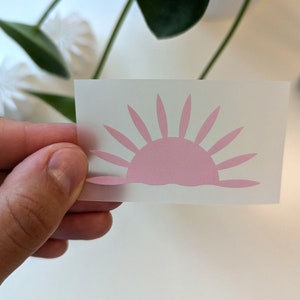 Könnte beinhalten: Ein rosa Vinyl-Aufkleber mit einer einfachen Silhouette eines Sonnenaufgangs. Die Sonne hat eine abgerundete Form mit Strahlen, die sich von oben nach unten erstrecken.