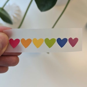 Könnte beinhalten: Eine Bogen mit Regenbogen-Herzaufklebern. Die Herzen sind in einer Reihe von links nach rechts angeordnet: rot, orange, gelb, grün, blau und pink.