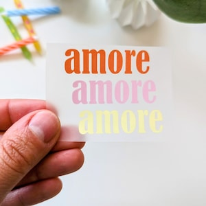 Könnte beinhalten: Ein transparenter Aufkleber mit dem Wort "amore" in Orange, Rosa und Gelb. Der Aufkleber wird vor einem weißen Hintergrund gehalten. Geburtstagskerzen und ein dekoratives Element sind ebenfalls sichtbar.
