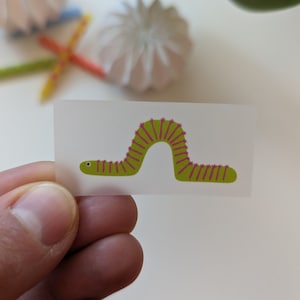 Könnte beinhalten: Ein kleiner, rechteckiger Aufkleber mit einer Cartoon-Raupe. Die Raupe ist grün mit rosa Akzenten und einem gebogenen Körper. Der Aufkleber wird in der Hand einer Person gehalten, mit anderen Objekten im Hintergrund.