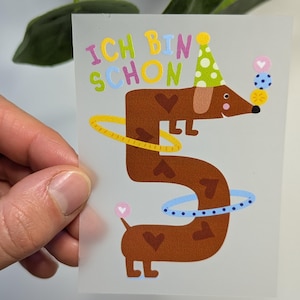 Könnte beinhalten: Eine farbenfrohe Illustration eines braunen Dackels, der die Zahl 5 bildet, mit Partyhut und bunten Objekten. Der Text "ICH BIN SCHON" steht darüber. Der Hund trägt einen gelben Reifen und einen blauen Reifen.