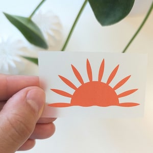 Könnte beinhalten: Eine weiße Karte mit einer einfachen orangenen Silhouette eines Sonnenaufgangs. Die Sonne hat eine abgerundete Form mit Strahlen, die sich von oben nach unten erstrecken.