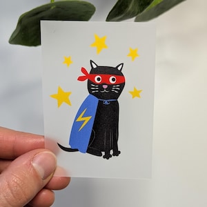 Könnte beinhalten: Eine schwarze Katze mit einer roten Maske und einem blauen Umhang mit einem Blitz-Design. Die Katze ist von gelben Sternen umgeben.