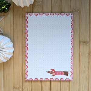 Könnte beinhalten: Notizblock mit rosa gewelltem Rand und gepunktetem Innenraum. Am unteren Rand befindet sich ein Cartoon-Dackel mit Weihnachtsmütze und Pullover. Dekorative Papierornamente und ein grüner Weihnachtsbaum aus Glas befinden sich im Hintergrund.