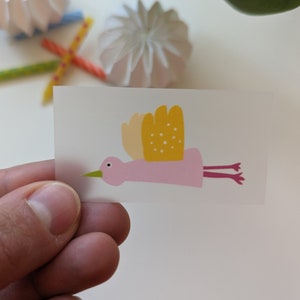 Könnte beinhalten: Ein rosa Vogel mit gelben Flügeln und weißen Punkten fliegt mit ausgestreckten Beinen. Der Vogel befindet sich auf einem durchsichtigen Aufkleber.
