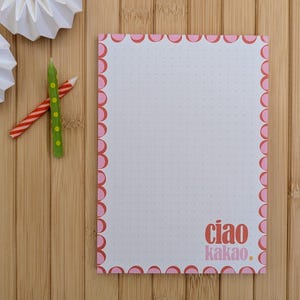Könnte beinhalten: Ein weißer Notizblock mit rosa Wellenrand und gepunktetem Rastermuster. Der Notizblock trägt den Text "ciao kakao." in Rosa. Zwei Origami-Papierdekorationen und zwei dekorative Kerzen auf einer Bambusoberfläche.