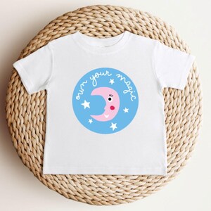 Könnte beinhalten: Weißes T-Shirt mit einem blauen Kreis-Design. Der Kreis enthält die Worte "own your magic" und einen rosa Mond mit Gesicht und Sternen. Das T-Shirt wird auf einer gewebten Korb-Oberfläche präsentiert.