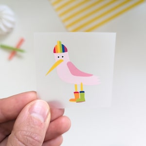 Könnte beinhalten: Ein Aufkleber mit einem Cartoon-Vogel mit rosa Körper, gelbem Schnabel und einem Regenbogenhut. Der Vogel trägt bunte Gummistiefel. Der Aufkleber wird von einer Hand gehalten.