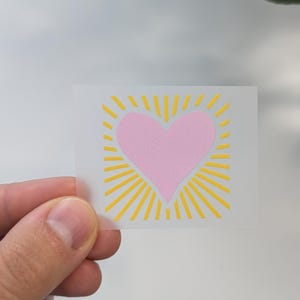 Könnte beinhalten: Ein pinkfarbenes Herz mit gelben Sonnenstrahlen, die nach außen strahlen. Das Herz befindet sich in der Mitte des Bildes und die Strahlen sind darum herum.