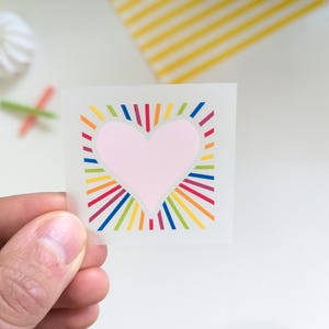 Könnte beinhalten: Ein quadratischer Aufkleber mit einem rosa Herzen in der Mitte, umgeben von farbenfrohen Regenbogenstrahlen. Der Aufkleber wird von einer Hand gehalten. Weitere Elemente im Hintergrund sind weiße Baisers und ein gelb-weiß gestreiftes Papier.