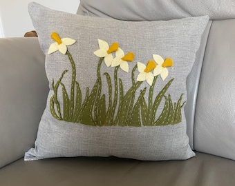 May include: A gray throw pillow with a white and yellow embroidered daffodil design. The daffodils are surrounded by green embroidered leaves.