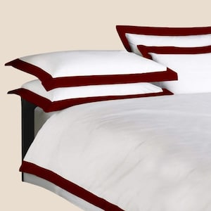Könnte beinhalten: Weißes Bettwäsche-Set mit einem dunkelroten Rand. Das Set enthält zwei Kissenbezüge und einen Bettbezug, alle mit einem sauberen, klaren Aussehen. Die Kissenbezüge sind auf dem Bettbezug gestapelt, wodurch ein geschichteter Effekt entsteht.