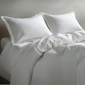 May include: A bed with white bedding, featuring two white pillowcases with a border. A white duvet cover is partially visible, creating a soft, inviting look. The bedding is neatly arranged, suggesting comfort and cleanliness. The overall aesthetic is simple and elegant.