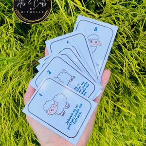 Puede incluir: Un montón de tarjetas de color azul claro con una ilustración de oveja de dibujos animados y texto en español. Las tarjetas están numeradas y tienen una parte superior redondeada. Las tarjetas se sostienen frente a un fondo de hojas verdes. El texto "Ats & Crafts by Michelle" está en un logotipo circular.