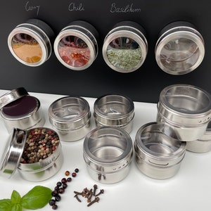 Könnte beinhalten: Satz Edelstahl-Gewürzgläser mit durchsichtigen Deckeln. Die Gläser sind magnetisch und können an einem Kühlschrank oder einer anderen Metalloberfläche befestigt werden. Die Gläser sind mit verschiedenen Gewürzen gefüllt, darunter Curry, Chili und Basilikum.