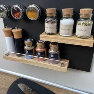Puede incluir: Un estante magn&eacute;tico para especias negro con dos estantes de madera. El estante superior tiene tres frascos de vidrio con tapas de corcho etiquetados como "Pfeffer", "Meersalz grob" y "Salatkr&auml;uter". El estante inferior tiene cuatro frascos de vidrio m&aacute;s peque&ntilde;os con tapas de corcho etiquetados como "Werkkraut No.1", "Werkkraut", "Werkkraut" y "Werkkraut".