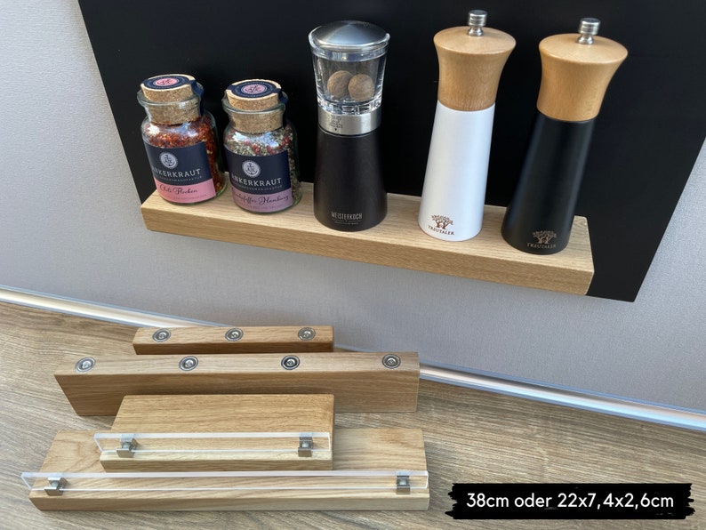 Puede incluir: Un juego de estantes magn&eacute;ticos para especias de madera con cuatro tarros de especias de vidrio y tres molinillos de pimienta de metal. Los tarros de especias est&aacute;n etiquetados como "Chili Powder" y "Greek Herbs". Los molinillos de pimienta son negros, blancos y de madera. Los estantes est&aacute;n montados en una pizarra negra.