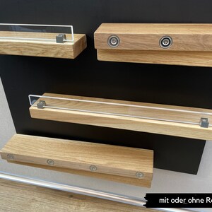 Puede incluir: Tres estantes de madera con cierres magn&eacute;ticos de metal. El estante superior tiene una barandilla de acr&iacute;lico transparente. Los estantes est&aacute;n montados en una superficie negra. El texto "mit oder ohne Reling" es visible en la parte inferior de la imagen.