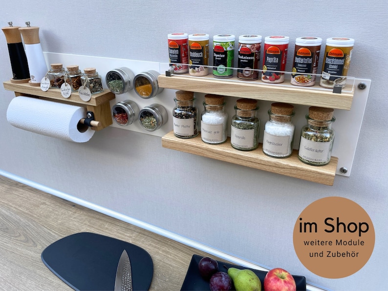 Puede incluir: Un estante para especias de pared blanco con dos estantes, un soporte para papel de cocina y tarros de especias magn&eacute;ticos. Los estantes son de madera y tienen un acabado marr&oacute;n claro. Los tarros de especias son de vidrio transparente con tapas de corcho y etiquetas. El estante est&aacute; montado en una pared blanca. El texto "im Shop" es visible en la esquina inferior derecha de la imagen. El texto "weitere Module und Zubeh&ouml;r" es visible debajo del texto "im Shop".