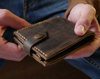 May include: A brown leather wallet with a flap closure and a brass button. The wallet is folded in half and has white stitching around the edges.