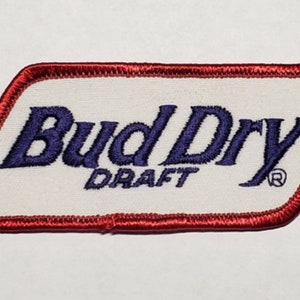 Könnte beinhalten: Ein weißer gestickter Aufnäher mit rotem Rand. Der Aufnäher zeigt den Text "Bud Dry Draft" in blauen Buchstaben mit einem eingetragenen Warenzeichen.