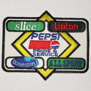 Pode incluir: Um remendo bordado branco, amarelo e preto com o logotipo da Pepsi e as palavras "Pride & Service". O remendo também apresenta os logotipos da Ocean Spray, Slice, Lipton e All Sport.