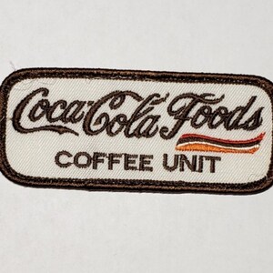 Może przedstawiać: Brązowa naszywka haftowana z białym tłem. Naszywka zawiera napis "Coca-Cola Foods Coffee Unit" z brązowo-pomarańczowym wzorem w paski.