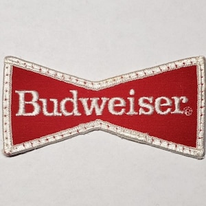 Könnte beinhalten: Ein fliegenförmiger Stickaufnäher mit rotem Hintergrund, dem weißen Schriftzug Budweiser und einem Symbol für eine eingetragene Marke. Der Aufnäher hat einen weißen gestickten Rand entlang des gesamten Umfangs der Stoffform.
